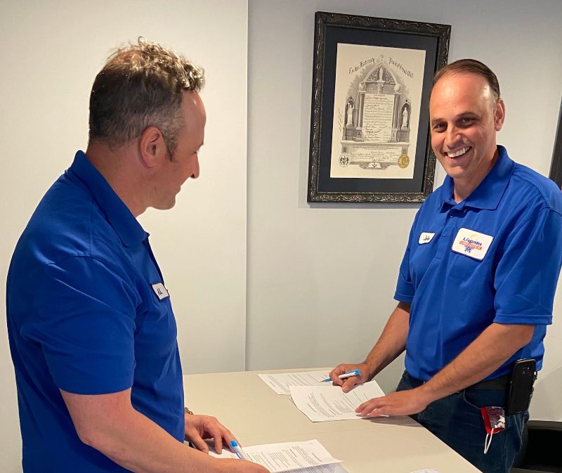 Al and his brother in their signature blue shirts are at a table with paperwork on it, signing the documents